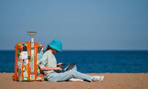 girl working on laptop while sitting against her suitcase on the beach
