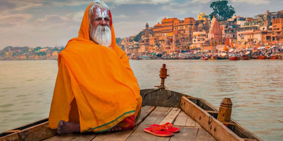 Sadhu baba in meditation on a wooden boat overlooking the historic Varanasi city and Ganges river ghats, India.