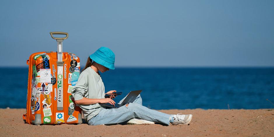 girl working on laptop while sitting against her suitcase on the beach
