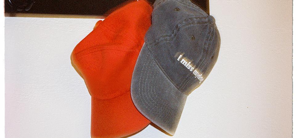 A pair of casual caps hang on a hook on a wall; one reads "I miss my dog"