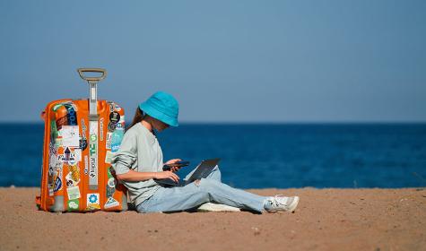 girl working on laptop while sitting against her suitcase on the beach