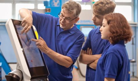 A teacher explains to two students, Gen Z trainees, how to operate a CNC machine.